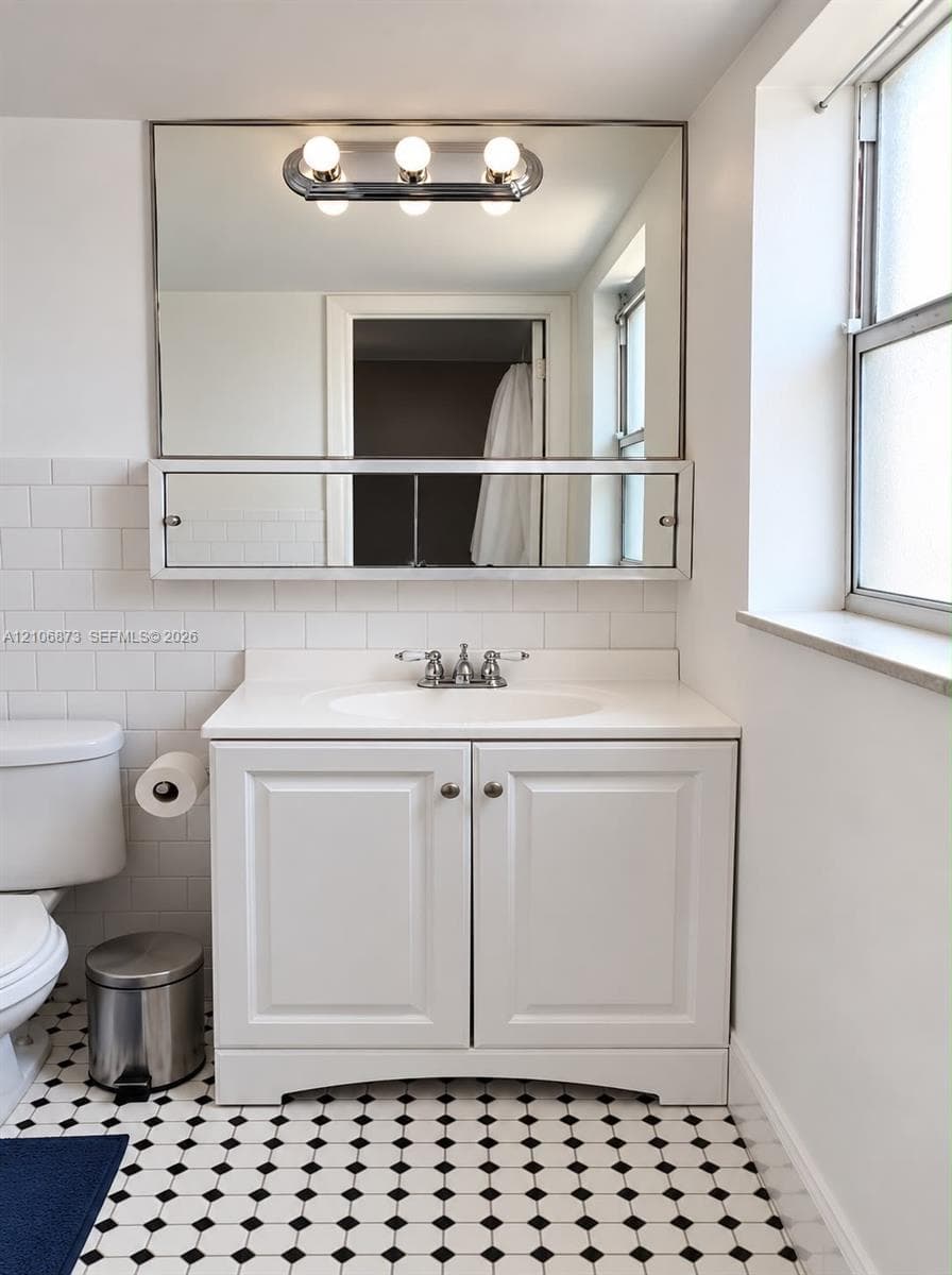 White vanity cabinet featuring a solid surface countertop with an integrated basin, wall-mounted medicine cabinet with mirror, three-light chrome vanity fixture, and patterned black-and-white tile flooring