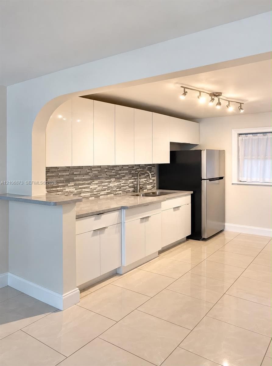 Kitchen featuring high-gloss white cabinetry, stone-finish countertops, decorative mosaic tile backsplash, stainless steel sink, and large-format tile flooring