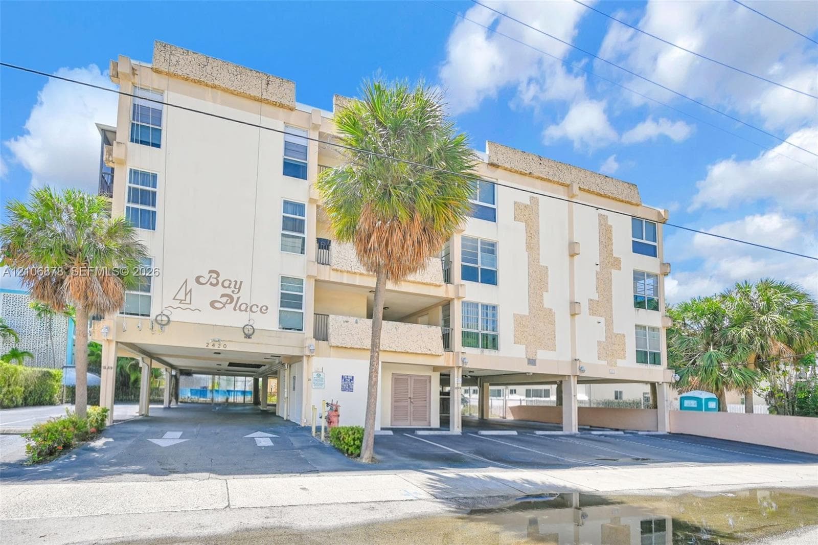 Multi-story residential building exterior featuring a stucco facade, textured stone accents, ground-level covered parking, and mature palm trees