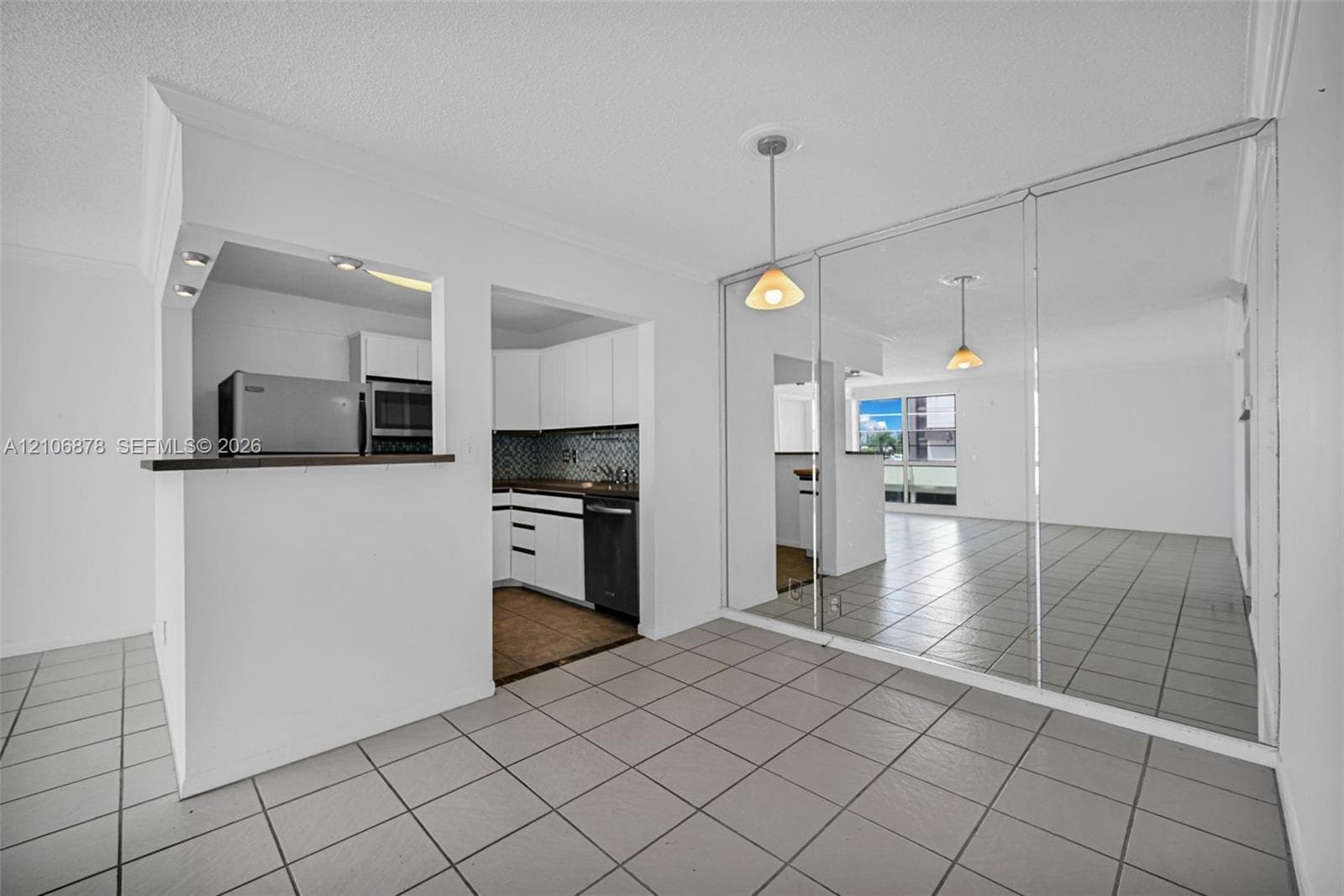 Kitchen featuring white cabinetry, dark countertops, tile backsplash, and a pass-through opening, finished with ceramic tile flooring and floor-to-ceiling wall-mounted mirrors