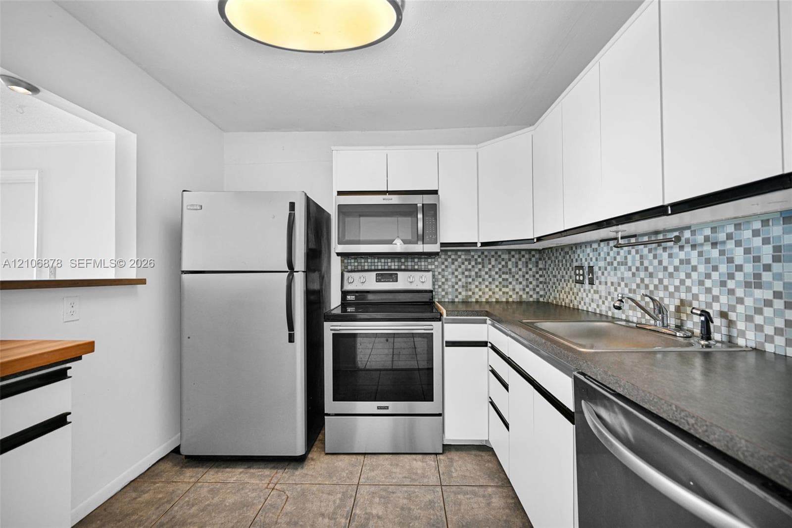Kitchen featuring white cabinetry, stainless steel appliances, multi-toned mosaic tile backsplash, dark countertops, and tile flooring