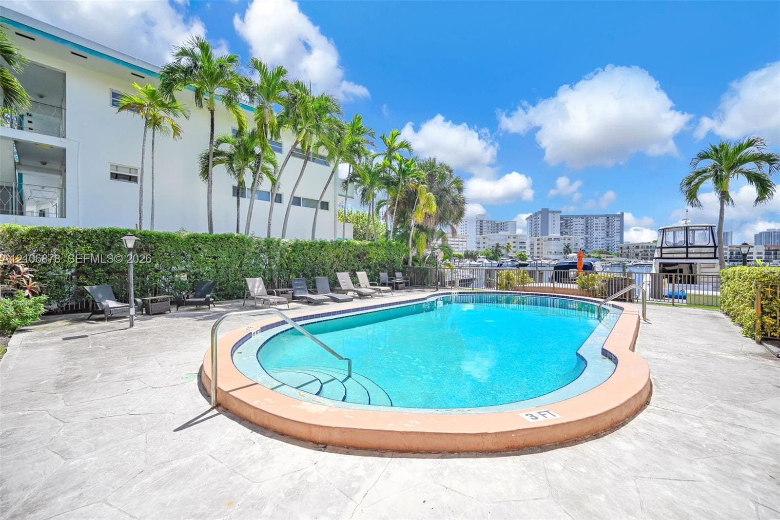 Outdoor swimming pool featuring a curved concrete deck, integrated entry steps, metal handrail, and surrounding mature palm trees