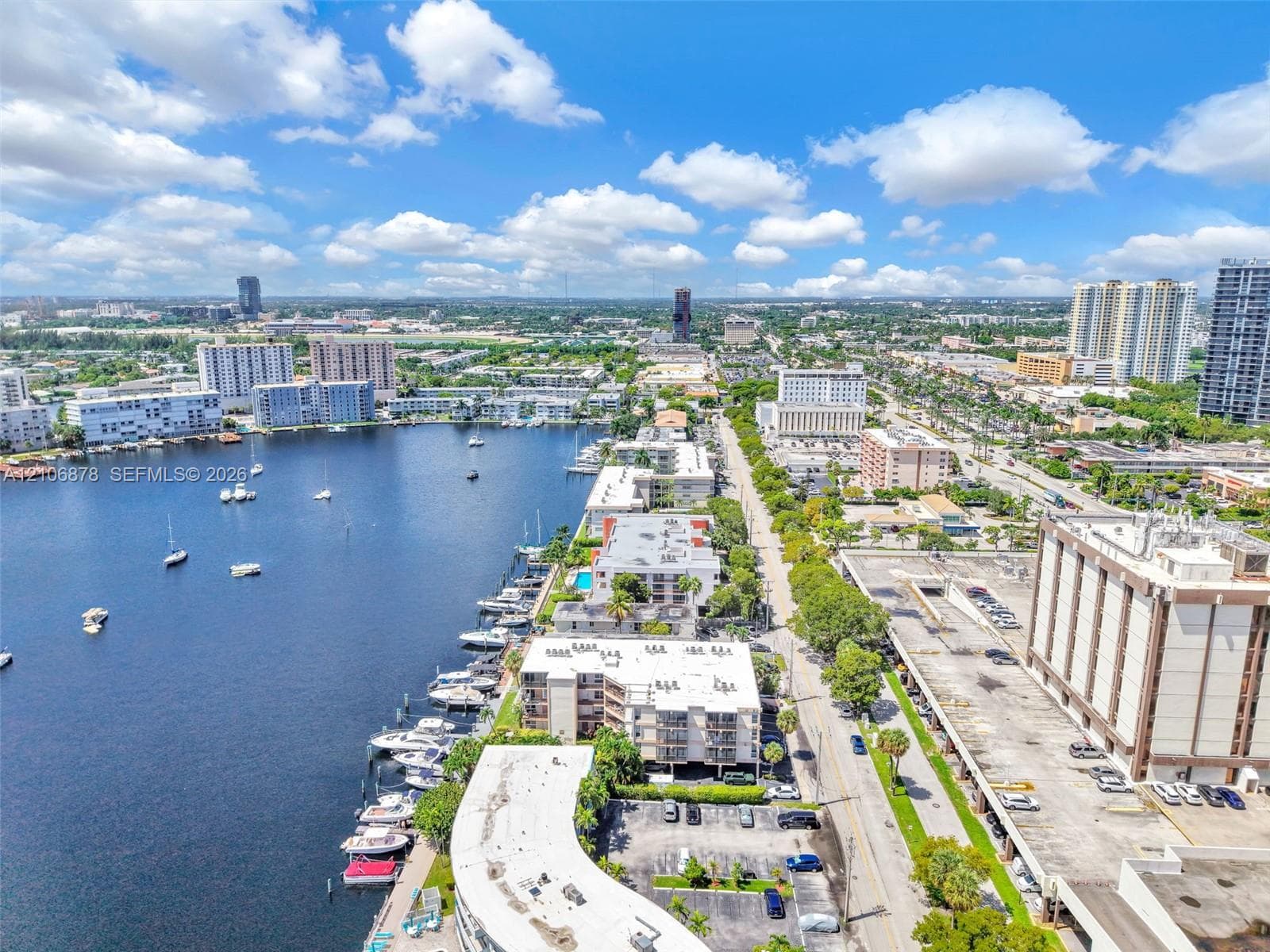 Aerial perspective of a waterfront setting featuring a canal, multi-level residential structures, surface parking, and urban infrastructure