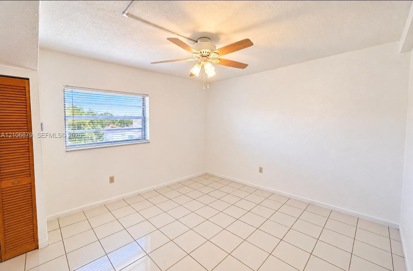 Neutral-toned interior space featuring light-colored tile flooring, a multi-light ceiling fan, a horizontal louvered closet door, and a single-pane window with aluminum horizontal blinds