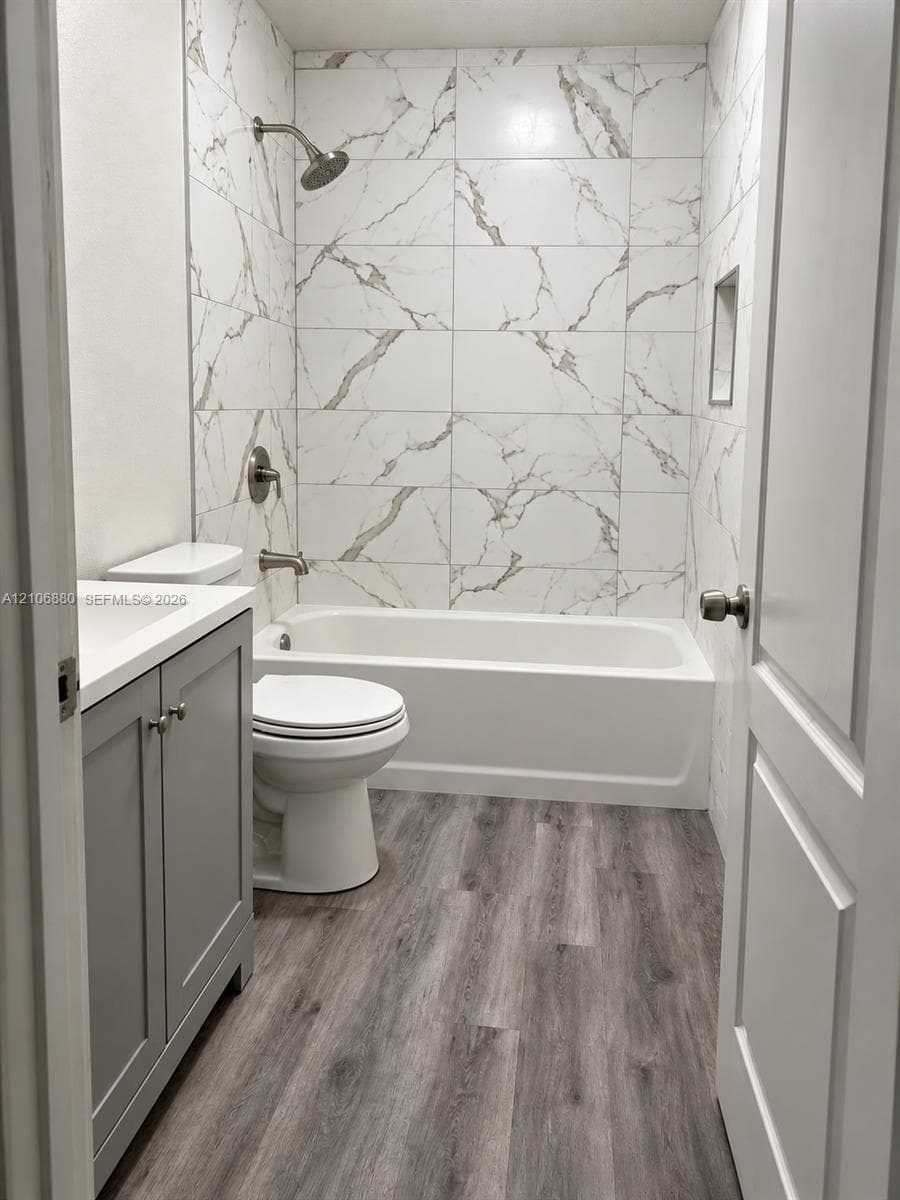 Bathroom featuring a vanity with a light-toned countertop, wood-finish flooring, and a tub-shower combination enclosed by large-format tile with decorative veining and a recessed shower niche
