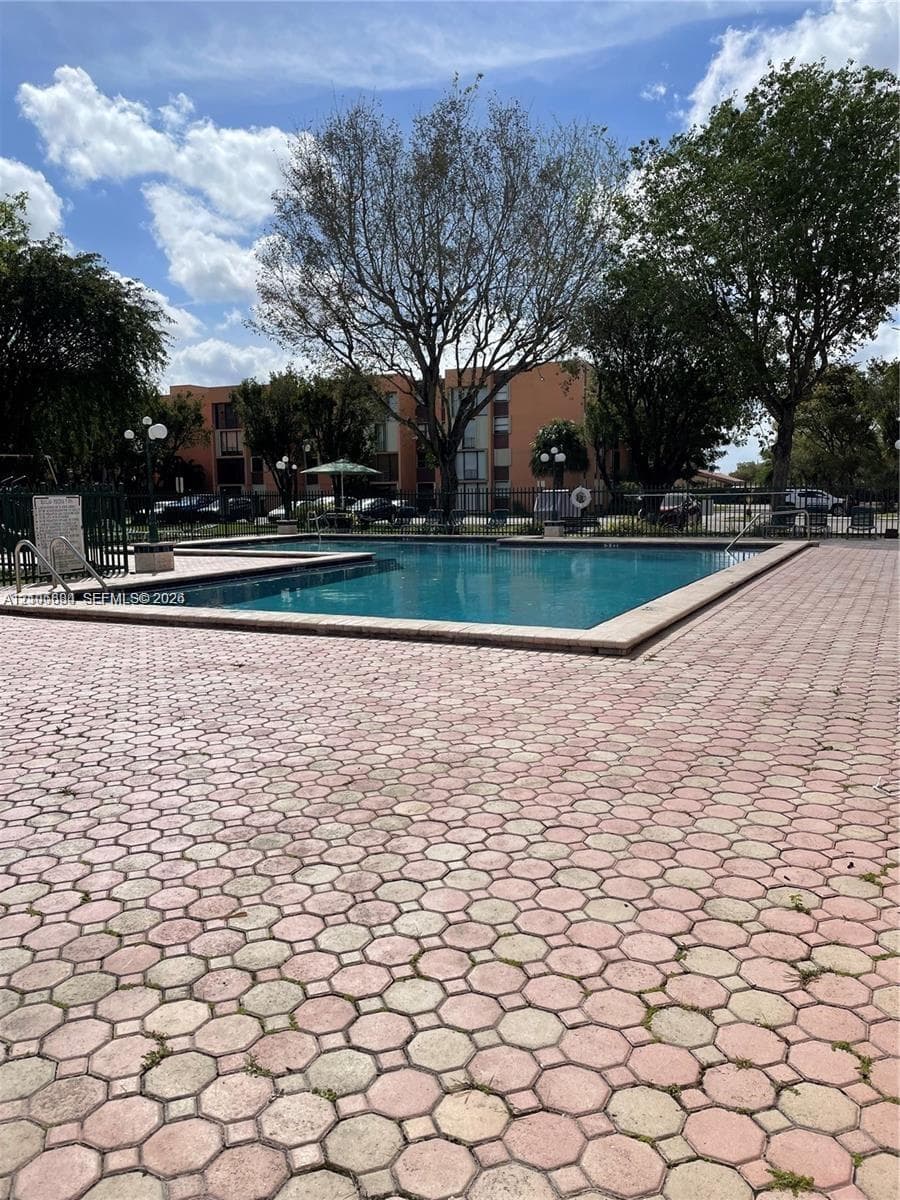 Outdoor swimming pool featuring a geometric paver deck, metal perimeter fencing, and mature tree canopy
