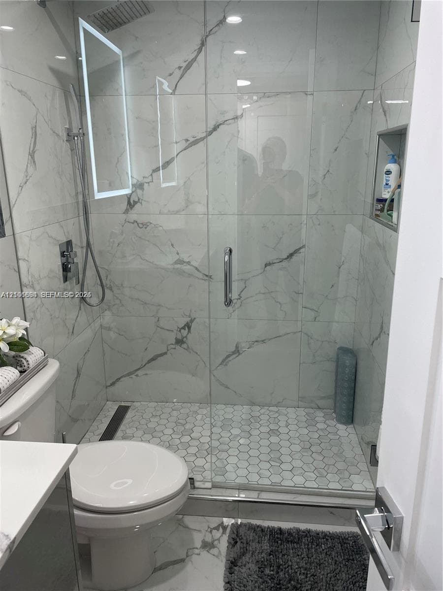 Walk-in shower featuring large-format veined tile surround, glass enclosure, recessed wall niche, and hexagon tile flooring, complemented by a wall-mounted rainfall shower head