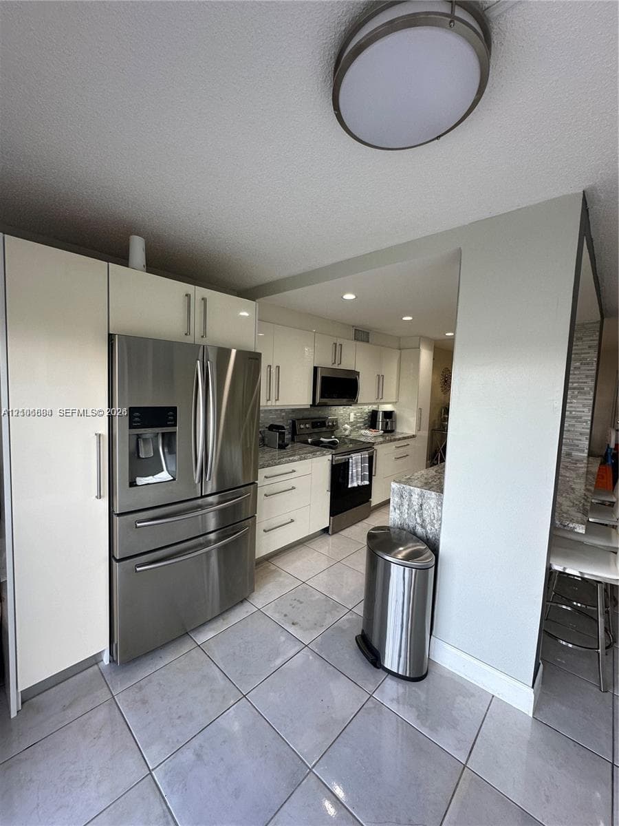 Kitchen featuring high-gloss white cabinetry, stone-finish countertops, stainless steel appliances, large-format tile flooring, and a flush-mount circular ceiling fixture