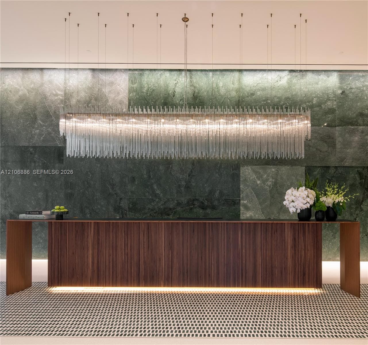 Large-scale linear chandelier with crystalline fringe, wood-veneer floating desk with base accent lighting, and full-height green-toned stone feature wall