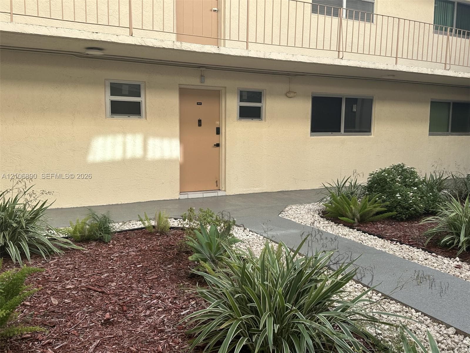 Stucco exterior featuring metal railings, sliding windows, and a paved walkway surrounded by landscaped mulch beds and stone borders