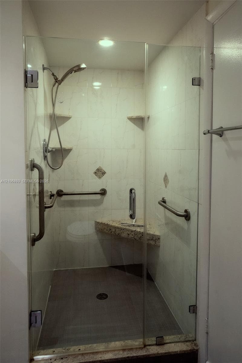Walk-in shower featuring a frameless glass enclosure, stone-surface corner bench, adjustable handheld showerhead, wall-mounted support rails, and tiled surround
