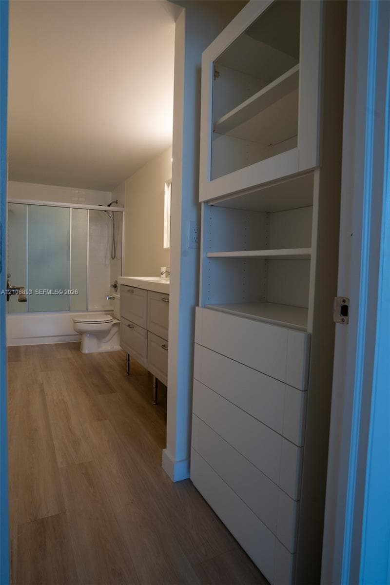 Built-in floor-to-ceiling white cabinetry featuring glass-fronted shelving and multiple drawers, wood-finish flooring, white vanity with rectangular basin, and a tub-shower combination with sliding frosted glass doors