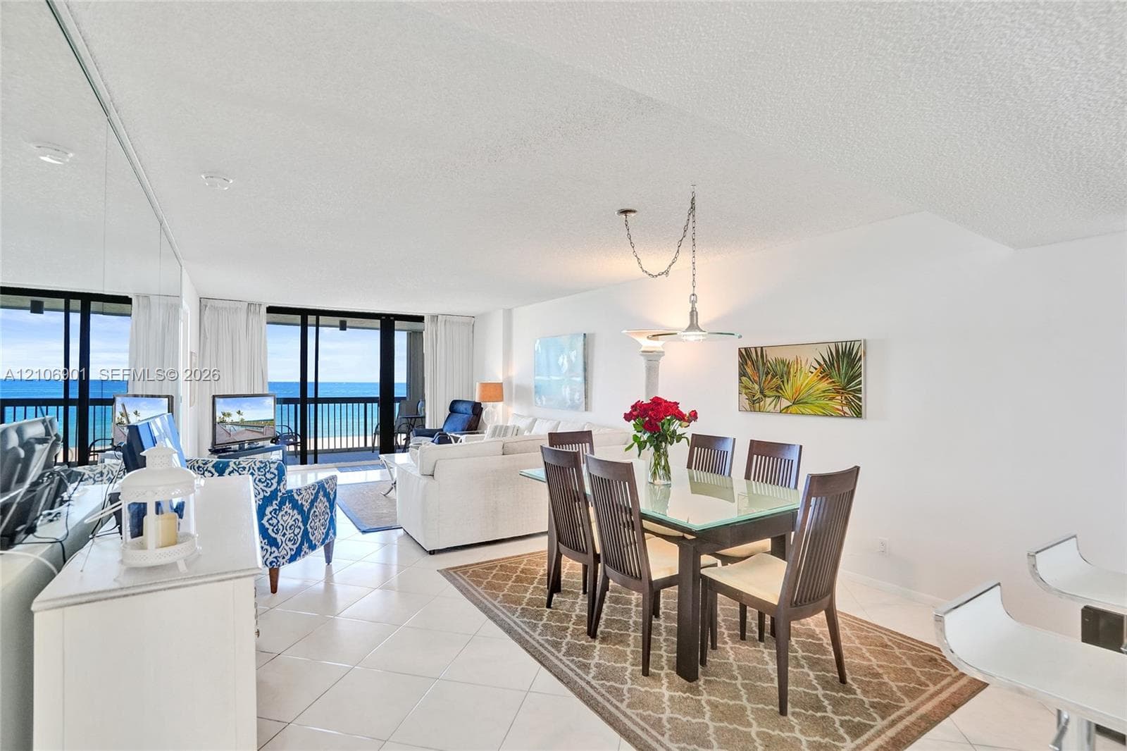 Open-concept interior featuring light-colored floor tiling, mirrored wall paneling, a tiered pendant light fixture, and floor-to-ceiling sliding glass doors opening to a balcony with ocean exposure