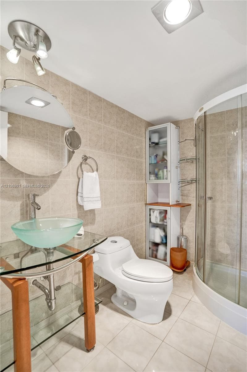 Glass vessel sink with chrome hardware, wood-frame pedestal vanity, curved glass-enclosed shower, built-in cabinetry, and ceramic tile wall and floor surfaces