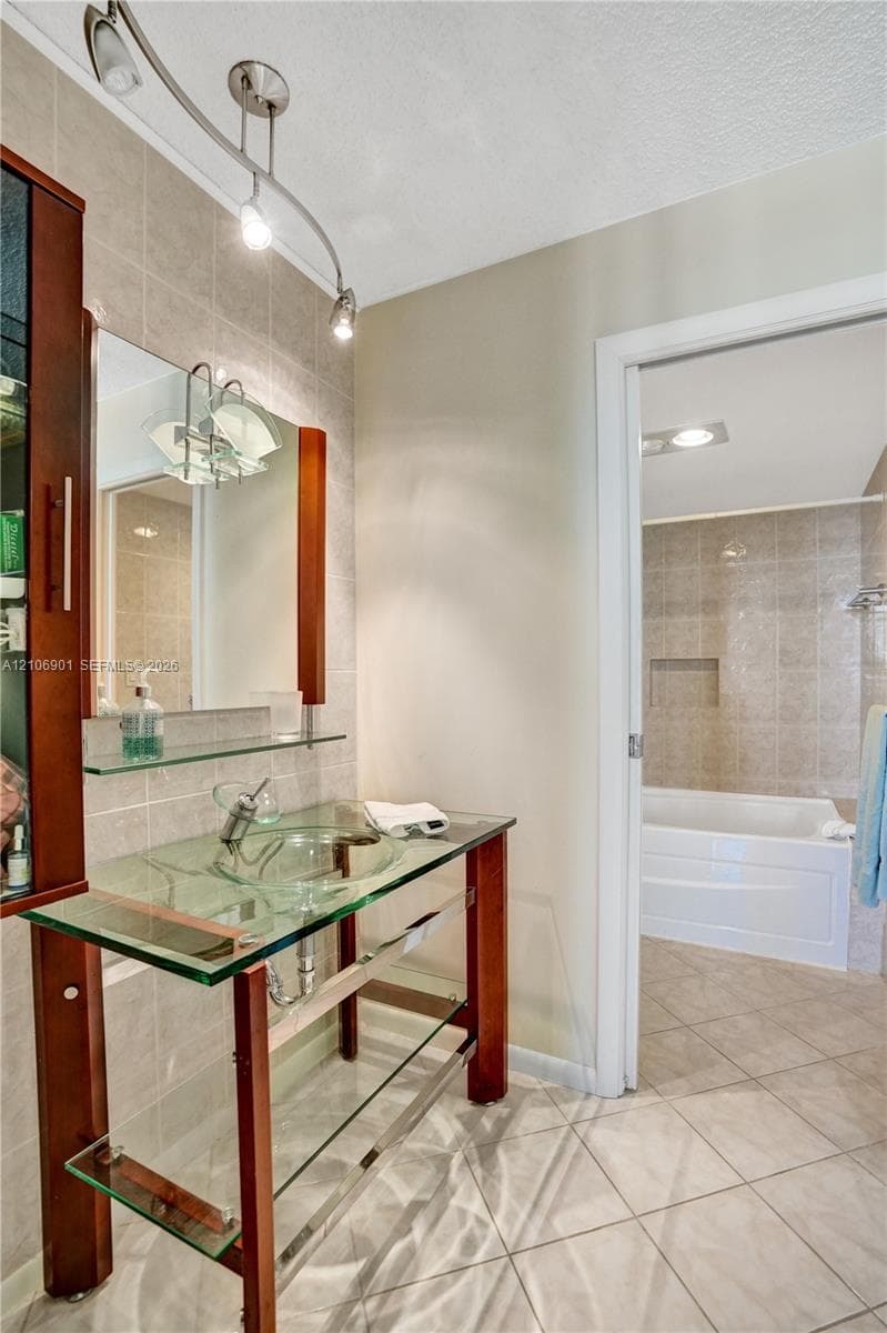 Glass vessel sink vanity featuring a wood-finish frame, clear glass shelving, wall-mounted track lighting, and a ceramic-tiled tub enclosure