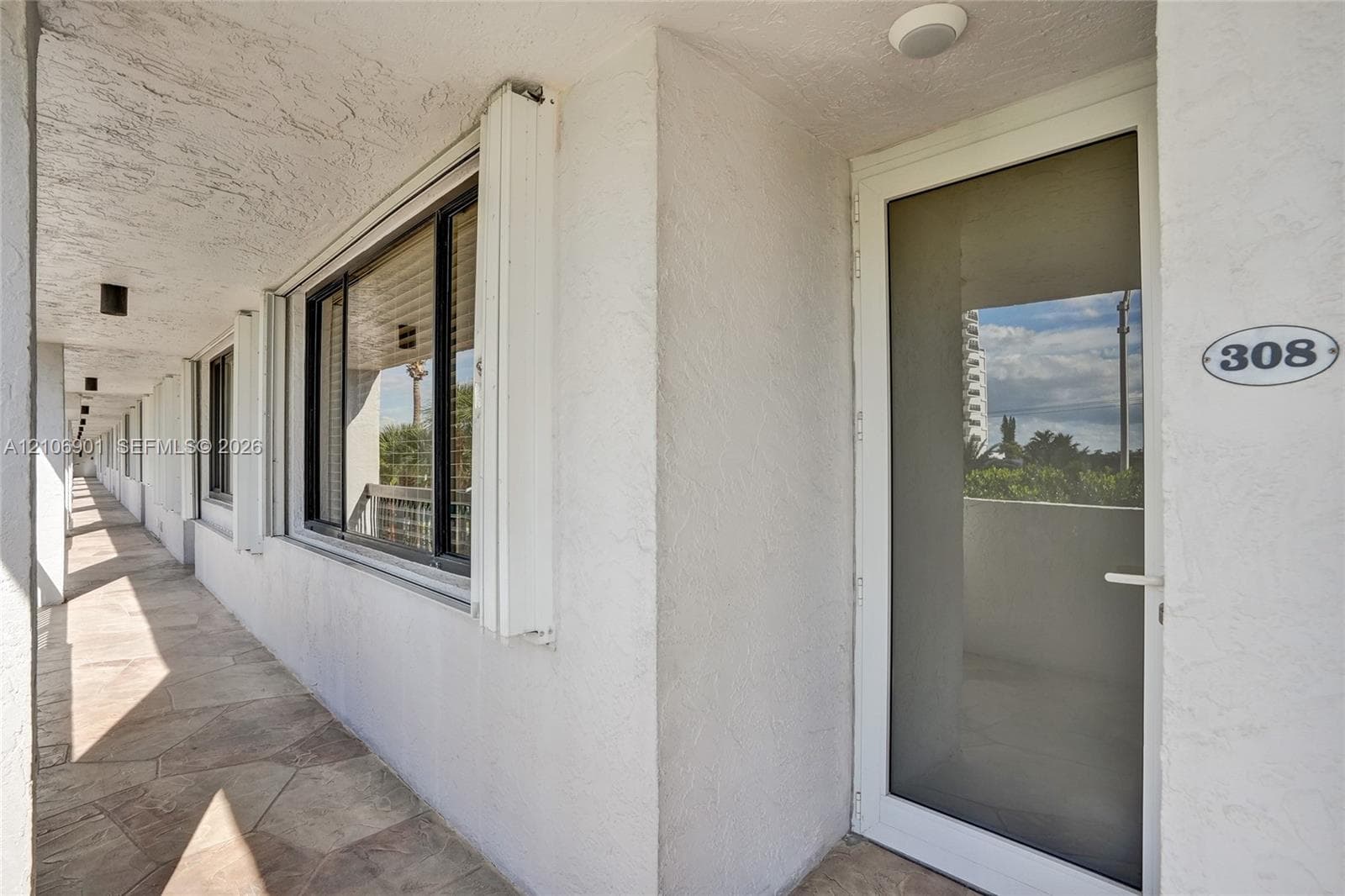 Exterior entry featuring a full-length glass storm door, textured white stucco walls, stone-finish tile flooring, and flush-mount exterior lighting