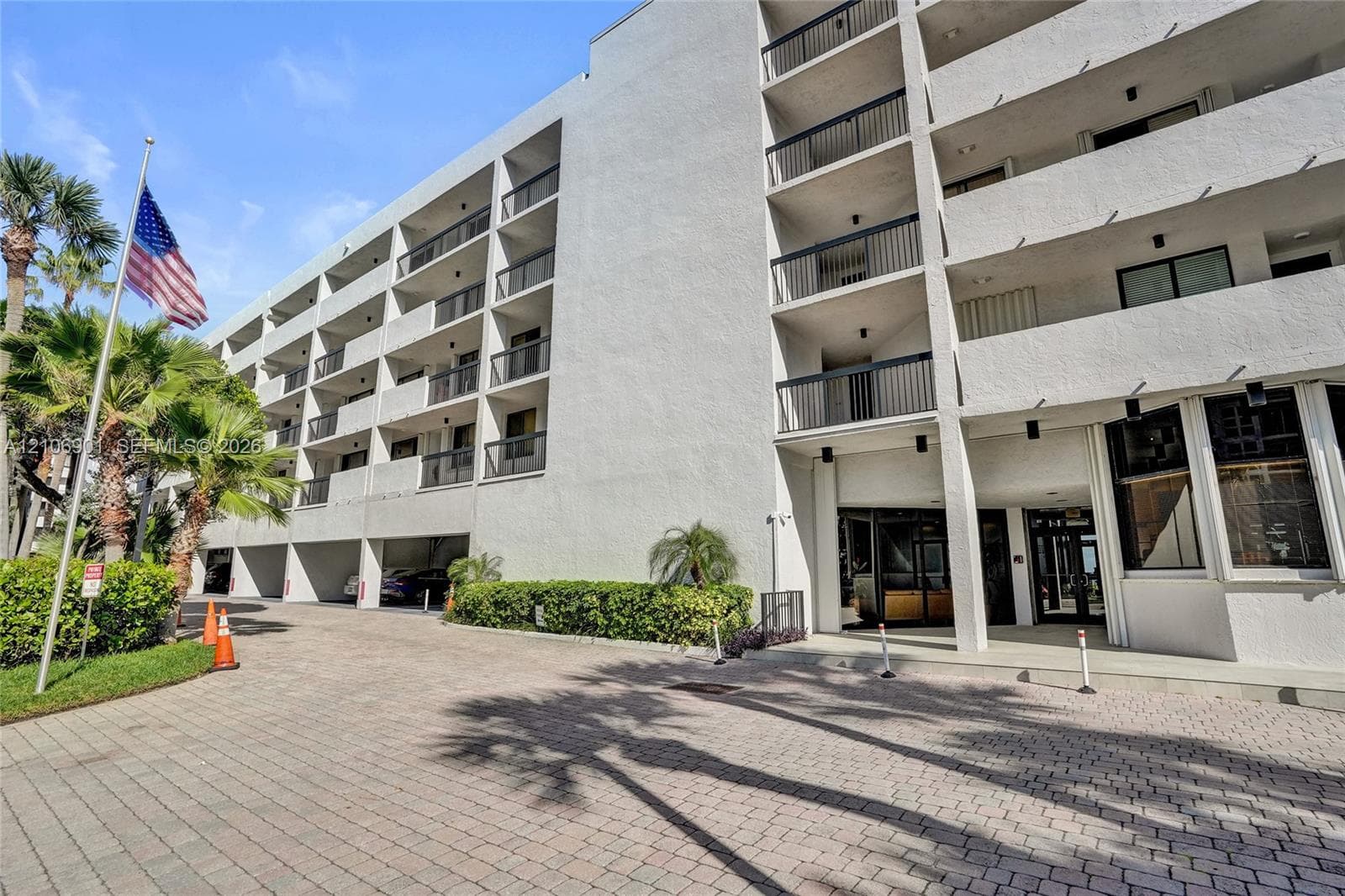 Multi-story concrete exterior featuring private balconies with black metal railings, ground-level covered parking, and paver stone driveway