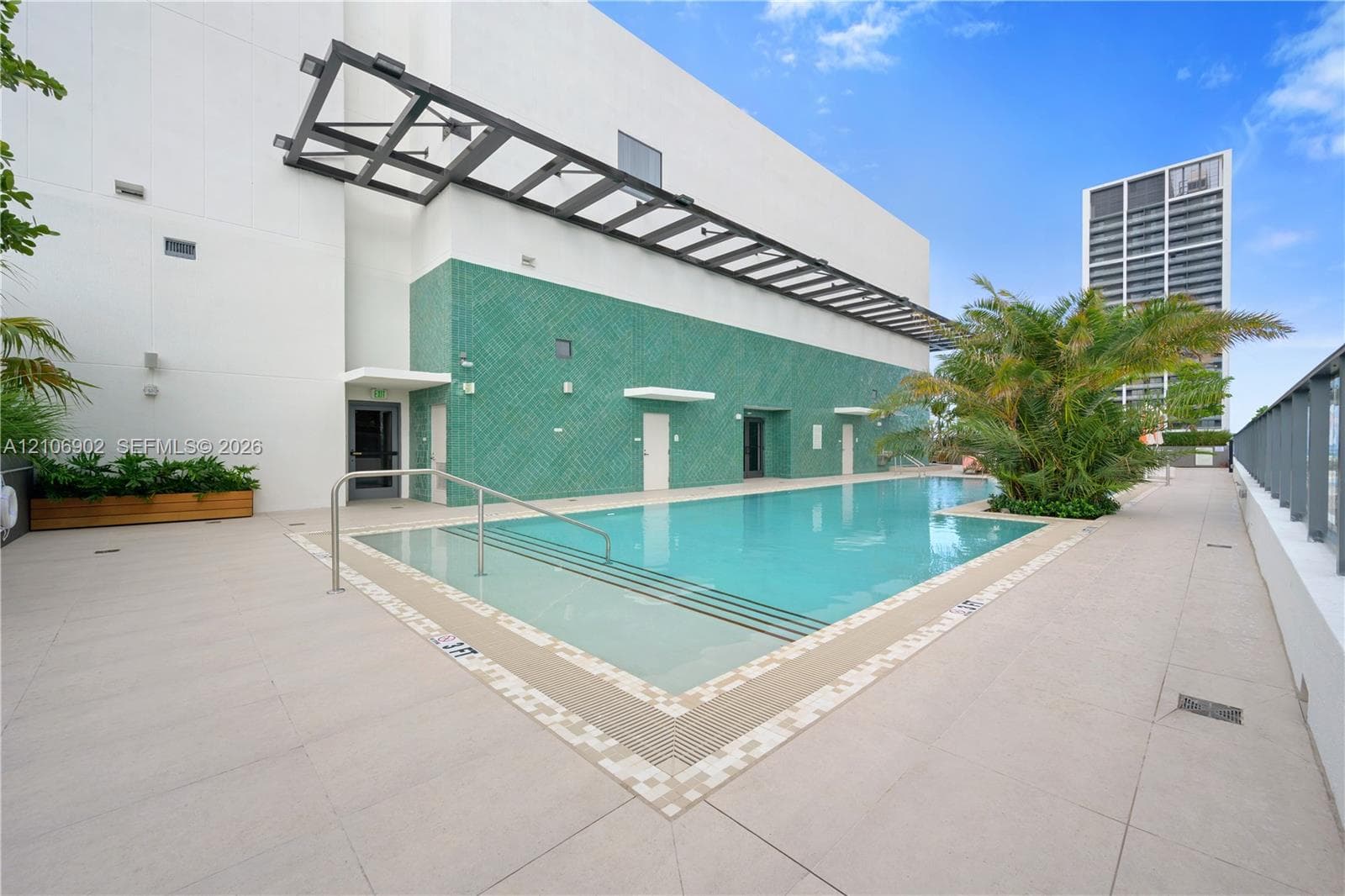 Swimming pool featuring a tiled surround, recessed entry steps, metallic handrail, and green textured accent wall with an overhead architectural pergola