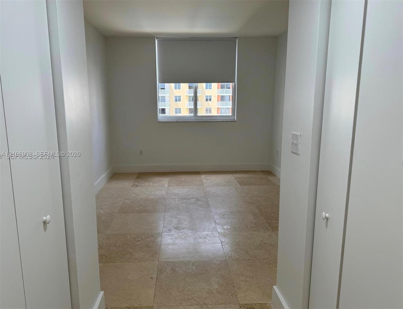 Neutral-toned tile flooring, dual closets with white swing doors, and a central window with a roller shade