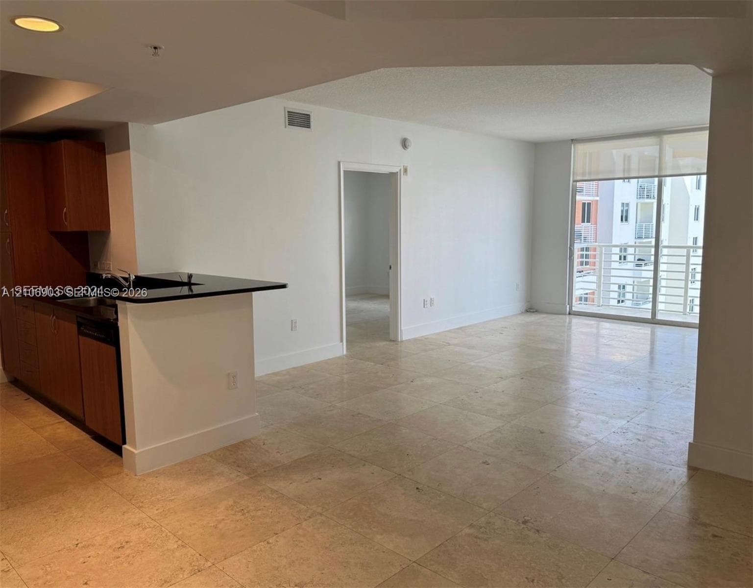 Open living area featuring large-format stone tile flooring, a kitchen peninsula with a dark solid-surface countertop, flat-panel wood-finish cabinetry, recessed ceiling lighting, and a glass sliding door providing balcony access
