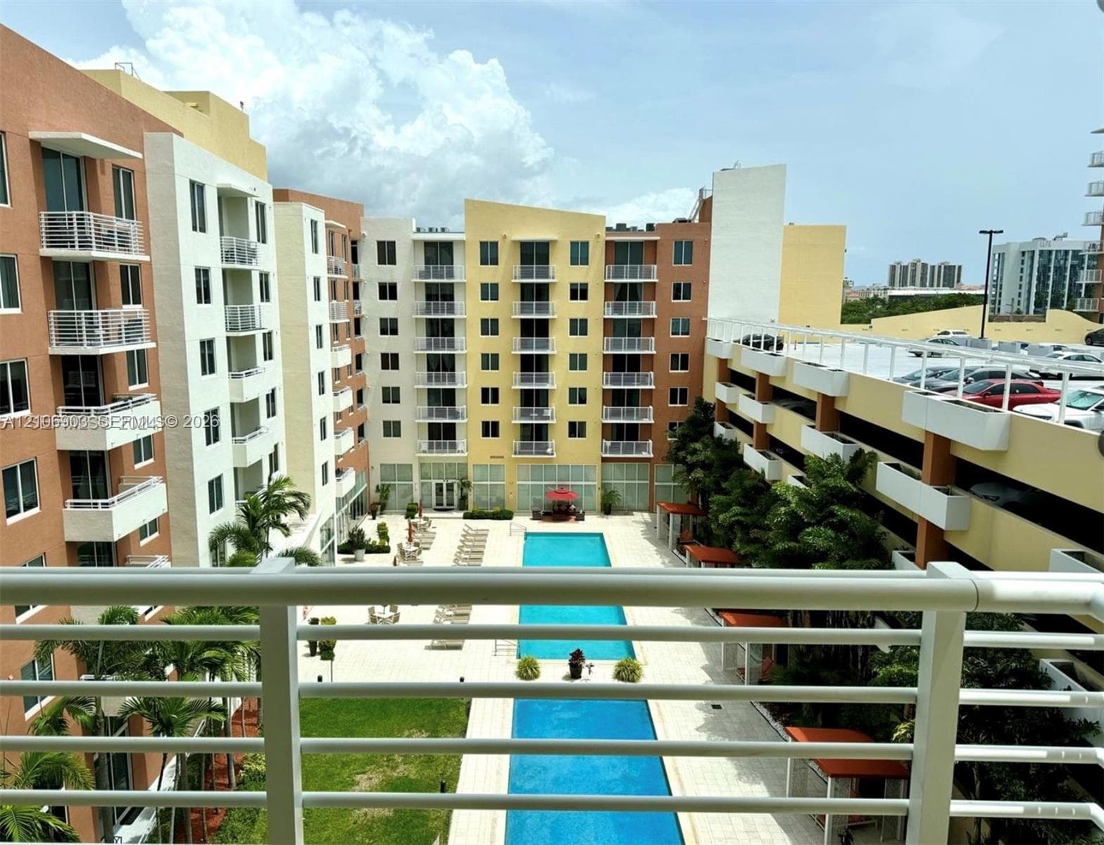 Multi-story residential complex featuring private balconies, a rectangular swimming pool, landscaped green space, and a structured parking facility