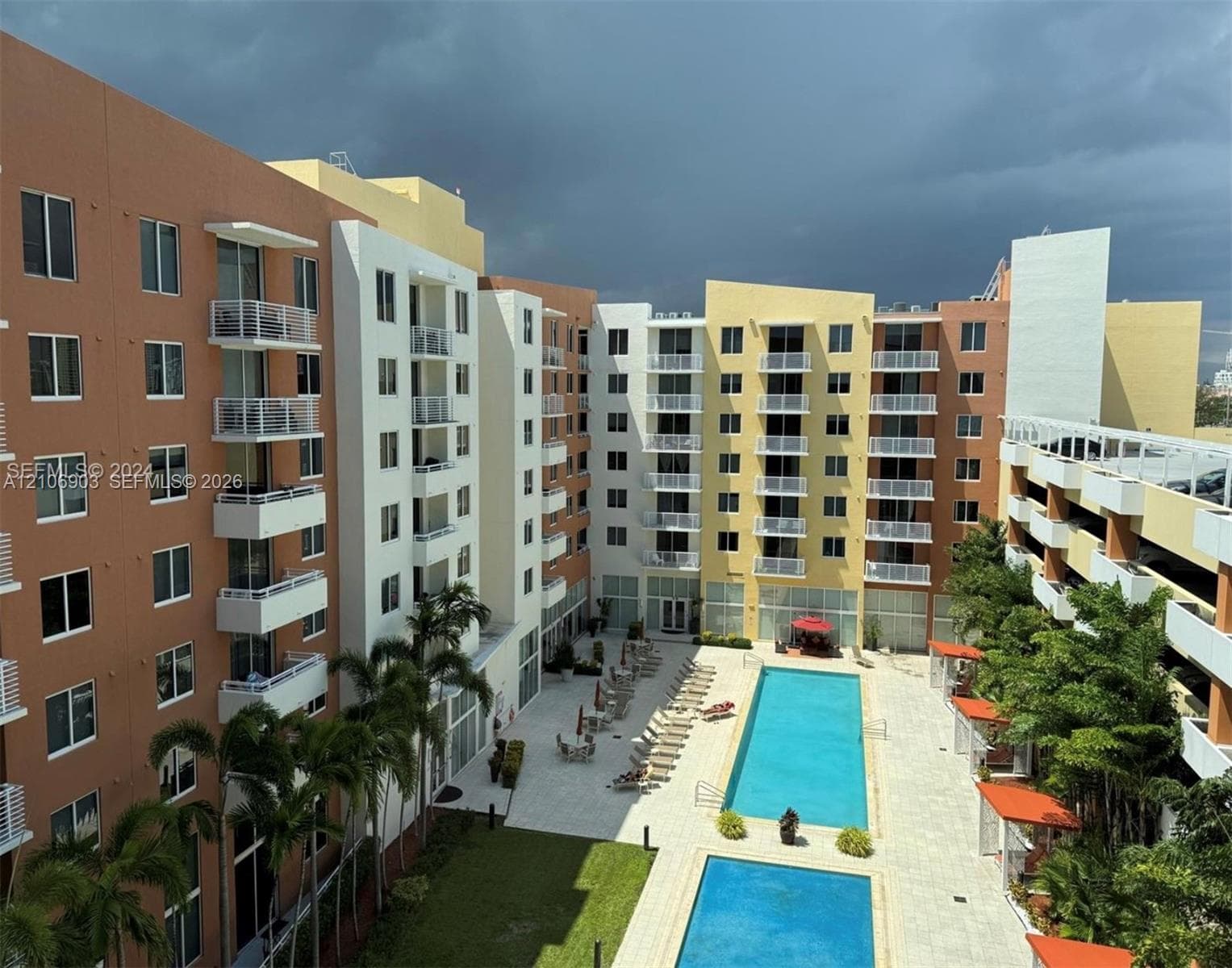 Multi-story residential complex featuring private balconies, dual outdoor swimming pools, stone-paved pool deck, and mature palm landscaping