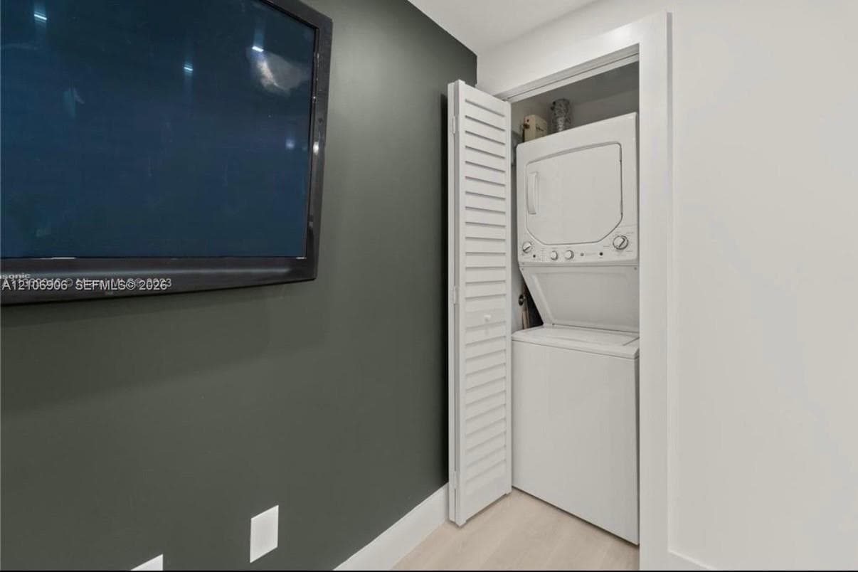 Stacked laundry unit housed within a closet featuring white louvered doors, contrasted by dark accent wall paint and light-toned flooring