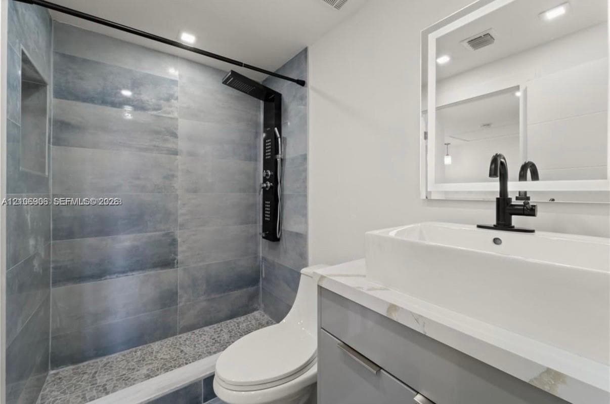 Bathroom featuring a walk-in shower with blue-toned large-format wall tile and a pebble-stone floor, a matte black shower panel system, a floating vanity with stone-finish countertop, a rectangular vessel sink with matte black faucet, and an integrated LED-backlit mirror