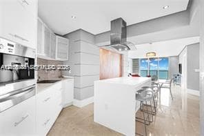 Modern kitchen featuring white flat-panel cabinetry, a central island with a solid-surface countertop, stainless steel vent hood, built-in coffee system, and polished tile flooring