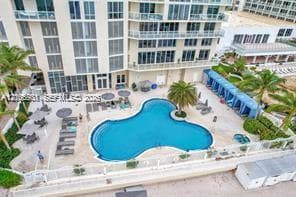 Multi-level building exterior featuring an organic-shaped swimming pool, paved pool deck, built-in cabanas, and mature palm trees