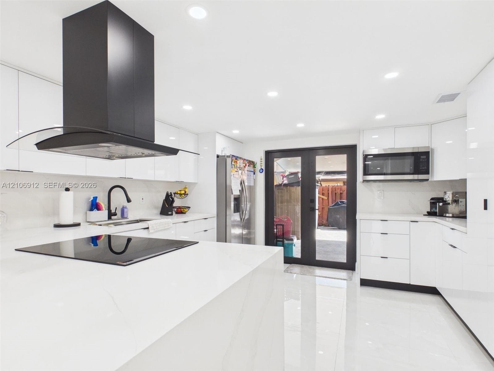 Modern kitchen featuring high-gloss white cabinetry, expansive stone countertops, a stainless steel built-in microwave, a matte black range hood, and glass-paneled double doors