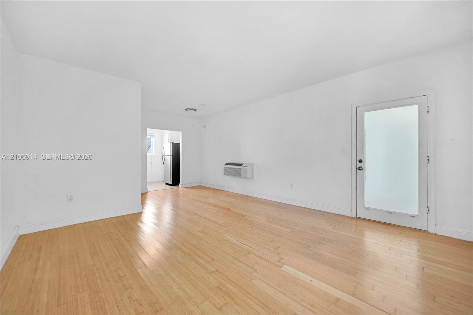 Open interior space featuring light wood-finish flooring, a wall-mounted climate control unit, and an exterior door with frosted glass paneling