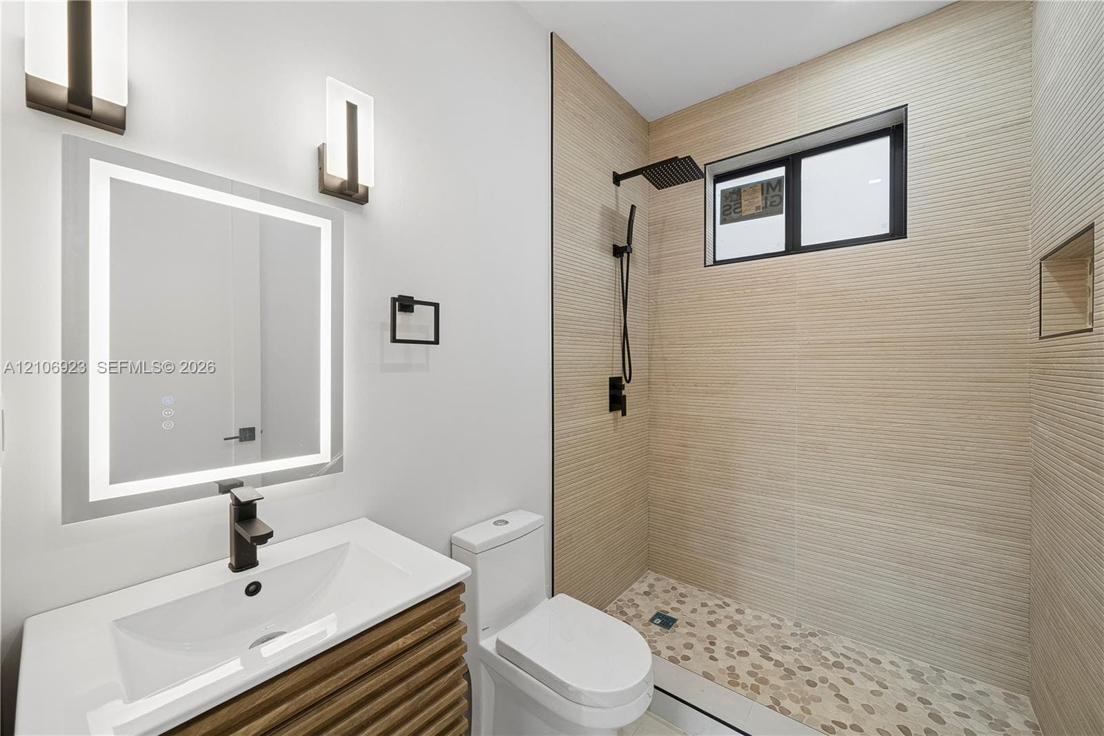 Bathroom featuring a vanity with wood-finish cabinetry, a lighted mirror, matte black wall-mounted faucet, walk-in shower with textured tile walls, and a pebble-stone floor