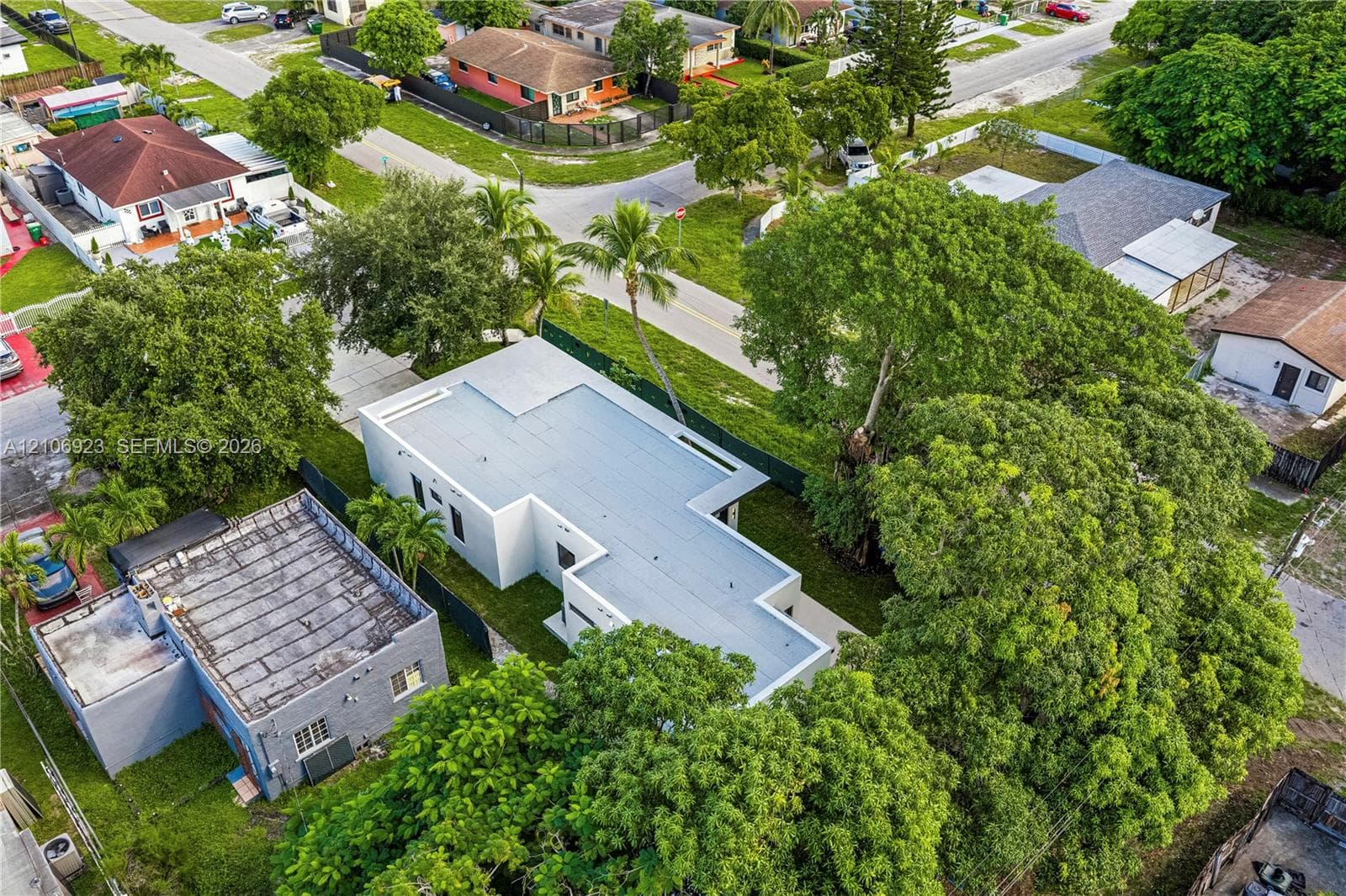 Aerial view of a rectangular property featuring a flat roof structure, concrete driveway, mature trees, and perimeter fencing