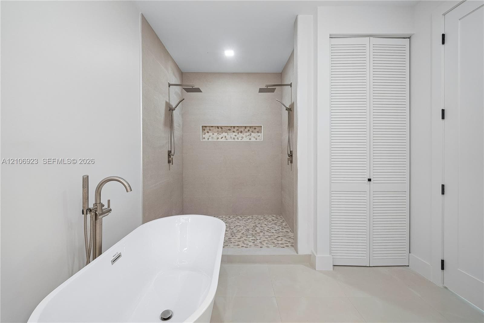 Freestanding soaking tub with metallic floor-mounted faucet, walk-in shower featuring dual overhead rainfall showerheads, recessed decorative tile niche, pebble-stone shower flooring, and louvered double-door cabinetry
