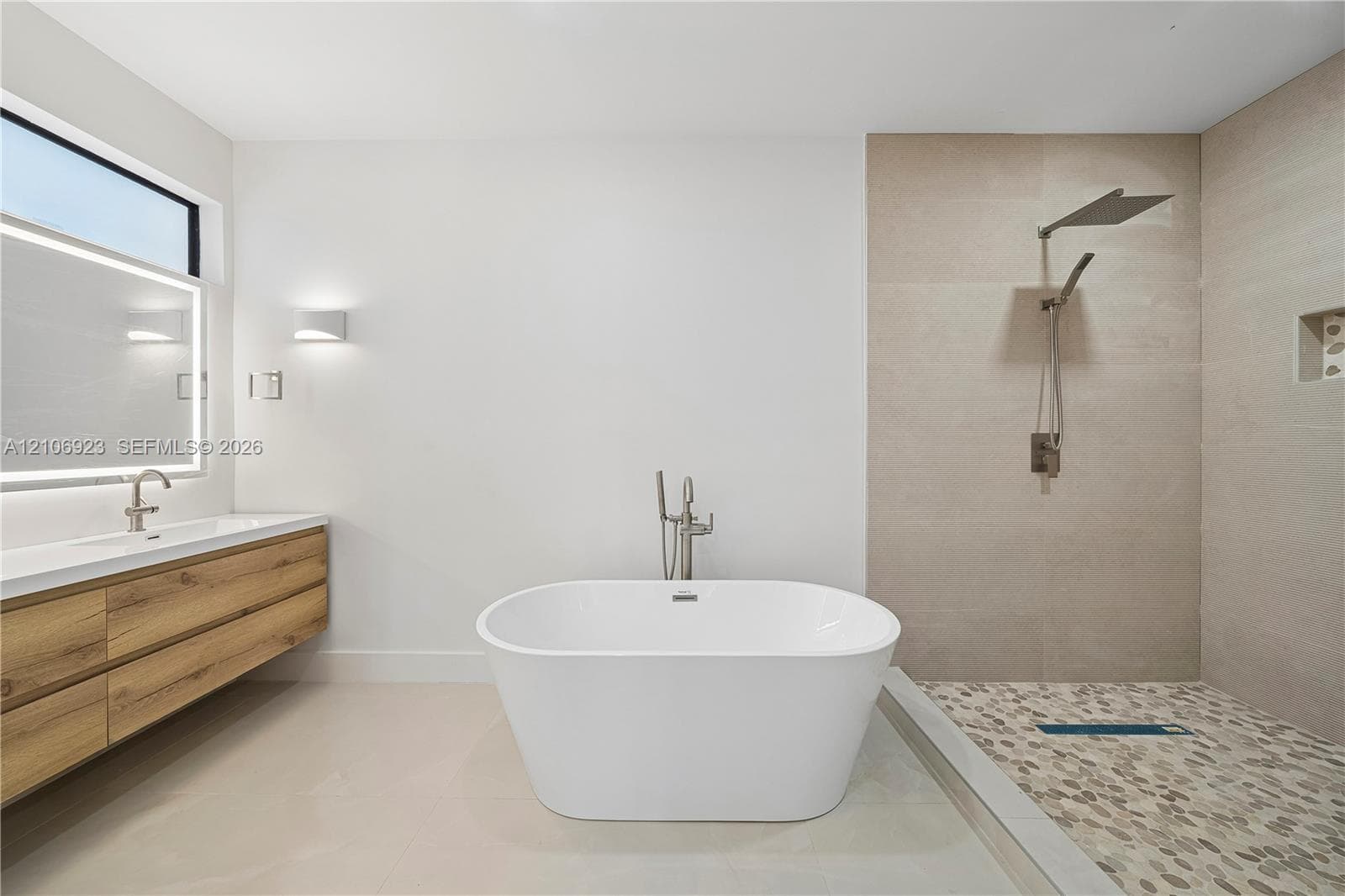 Freestanding soaking tub with floor-mounted faucet, floating wood-finish vanity, backlit mirror, walk-in shower with pebble-stone flooring, and wall-mounted rainfall showerhead