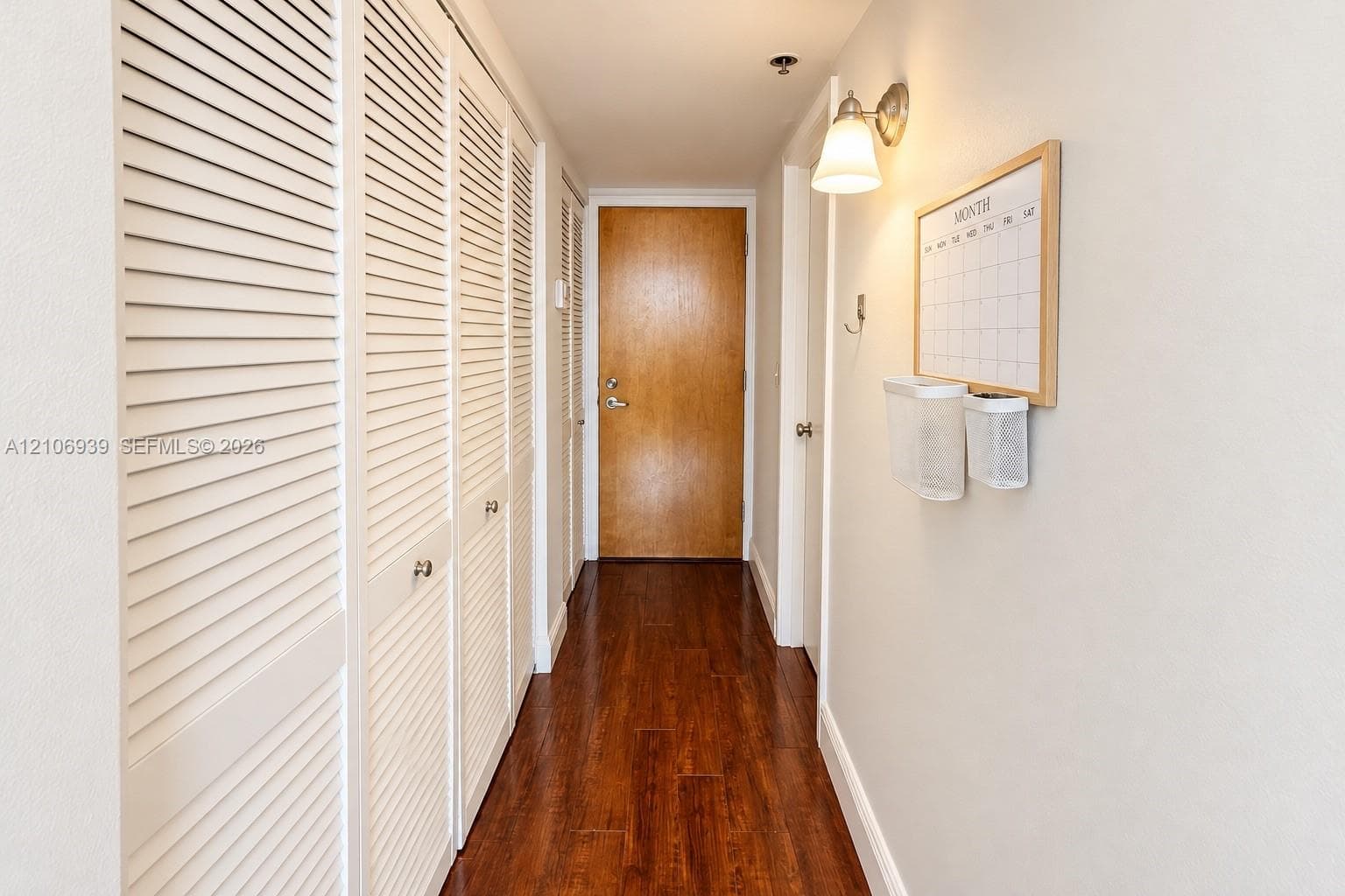 Hallway featuring wood-finish flooring, white louvered closet doors, and wall-mounted light fixture