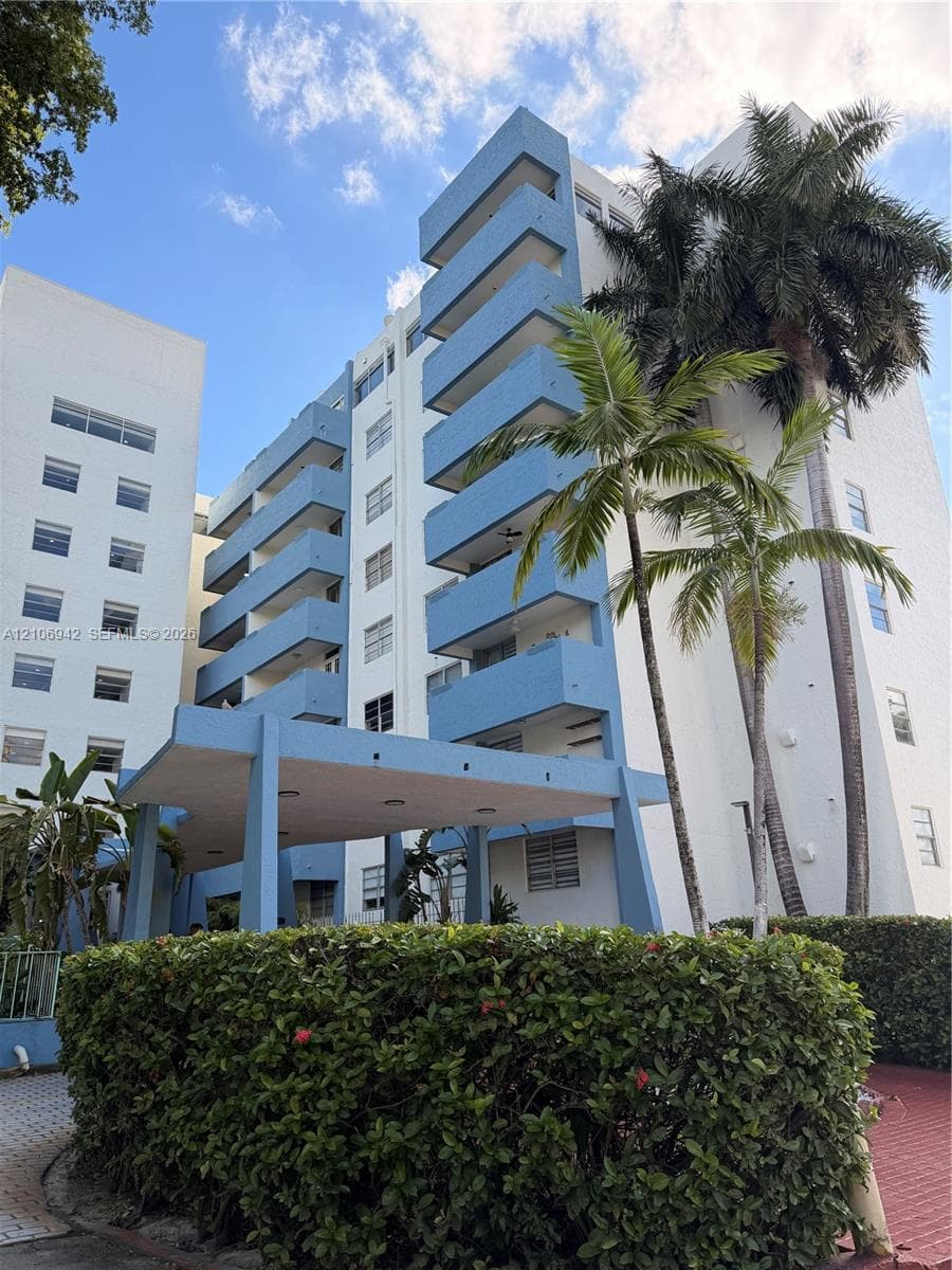 Multi-story residential building exterior featuring stacked private balconies, a structural covered entryway, and mature palm trees