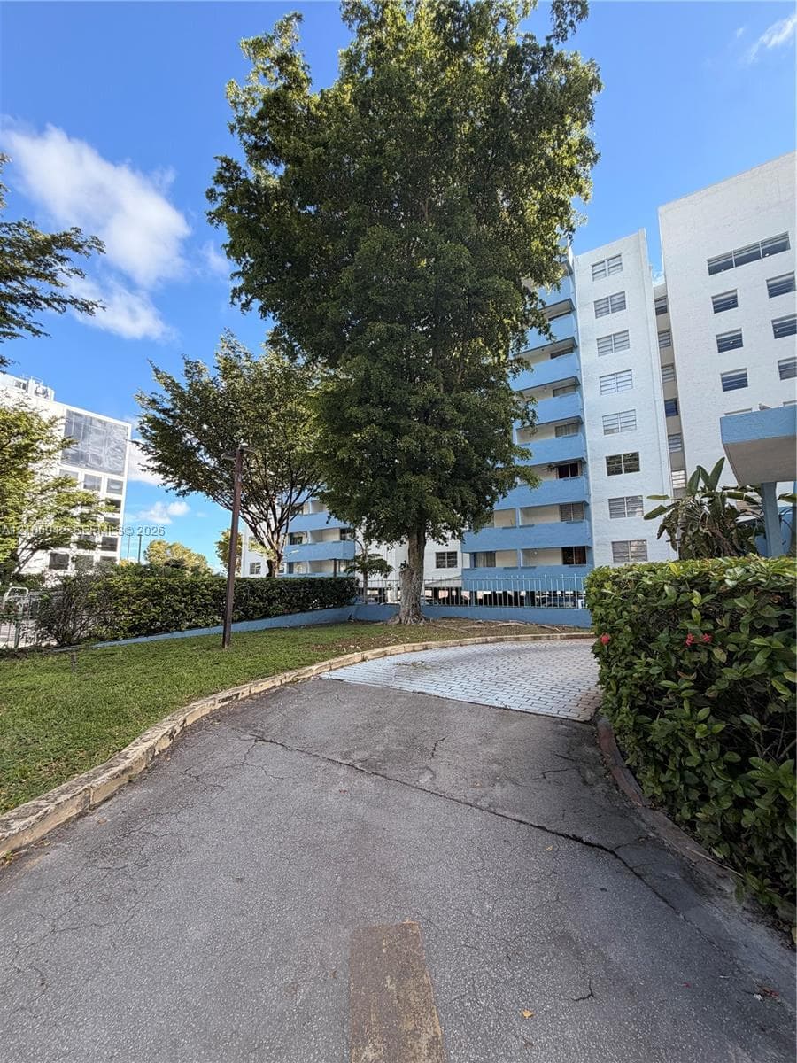 Exterior grounds featuring paved access, mature trees, manicured hedges, and multi-story building architecture