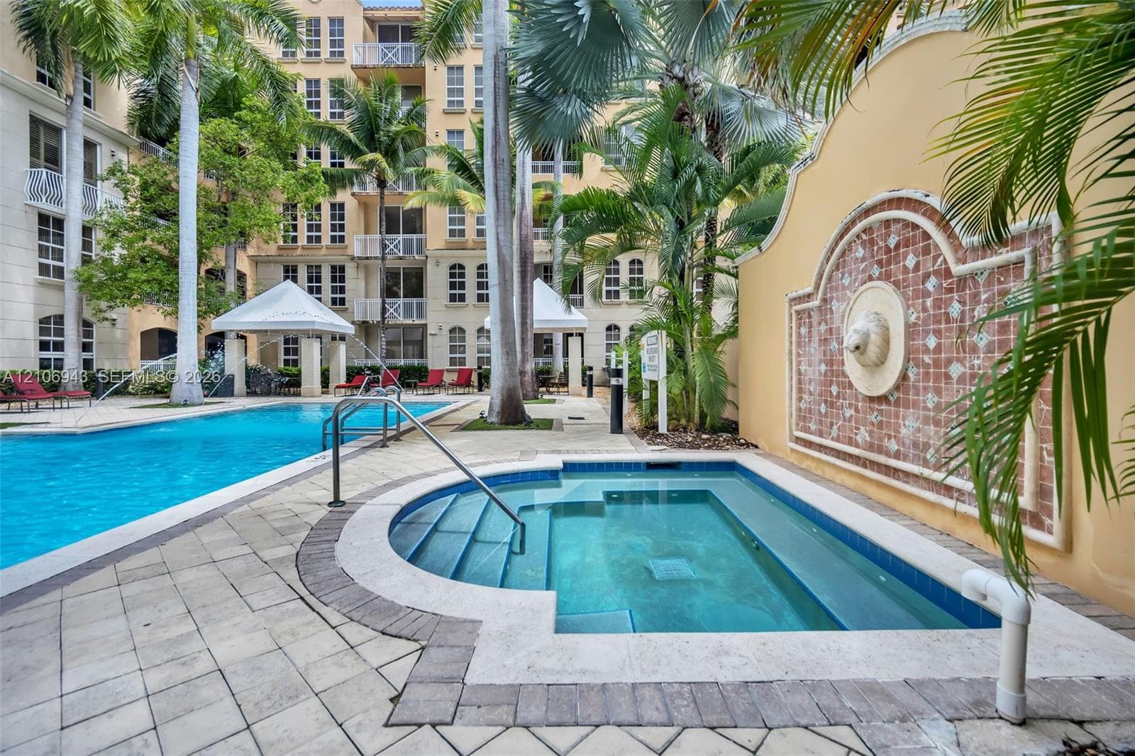 In-ground spa with stone coping, swimming pool, decorative tile wall fountain, stone-paver hardscaping, and mature palm trees
