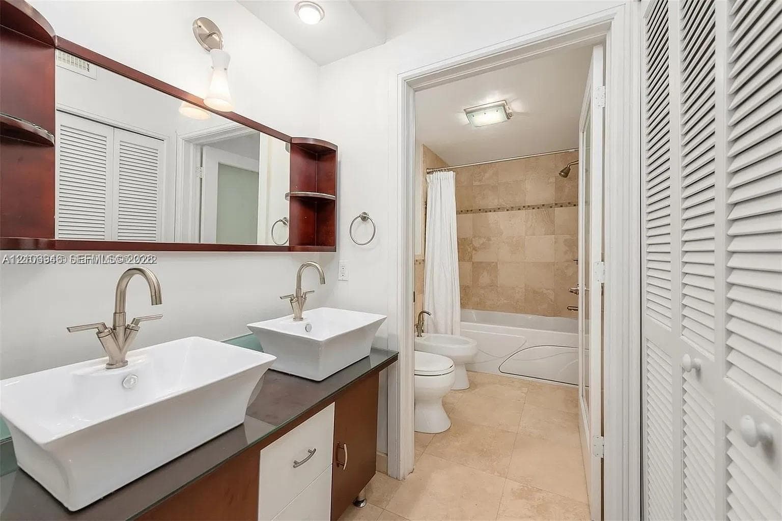 Dual vessel sinks with metallic finish faucets, dark-toned cabinetry, a built-in wood-frame mirror, stone-finish tile flooring, and a shower-tub combination with stone-style wall tile