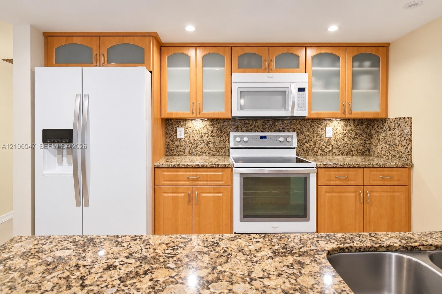 Kitchen featuring custom wood cabinetry with glass-panel upper doors, stone-finish countertops, stone-finish backsplash, and built-in white appliances