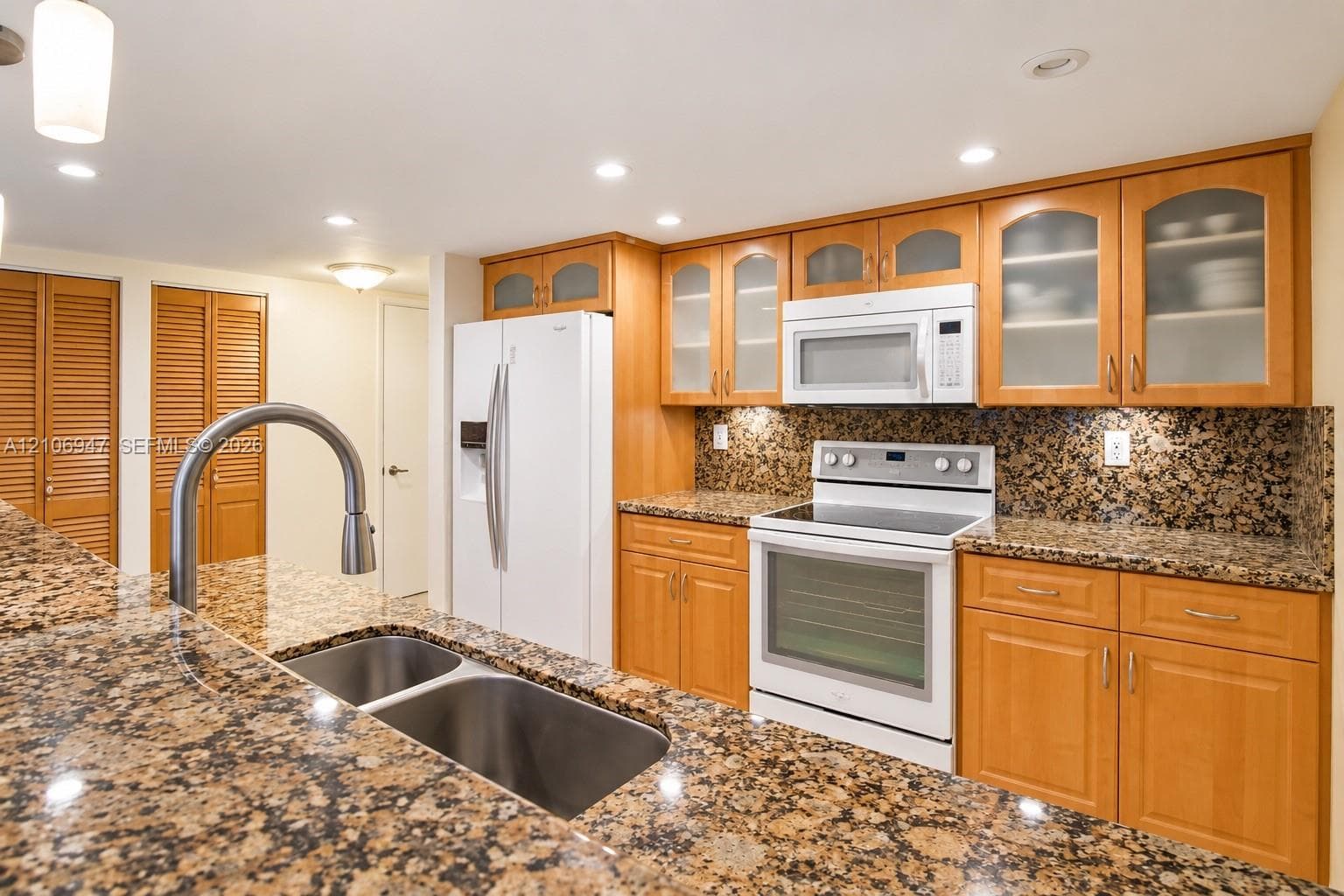 Kitchen featuring natural wood-finish cabinetry, stone countertops, double-basin stainless steel sink with high-arc faucet, and upper glass-front display cabinets