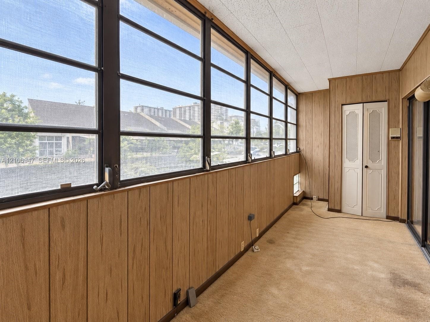 Enclosed space featuring vertical wood-finish wall paneling, a wall of multi-pane windows with metal framing, textured ceiling tiles, and dual-panel closet doors