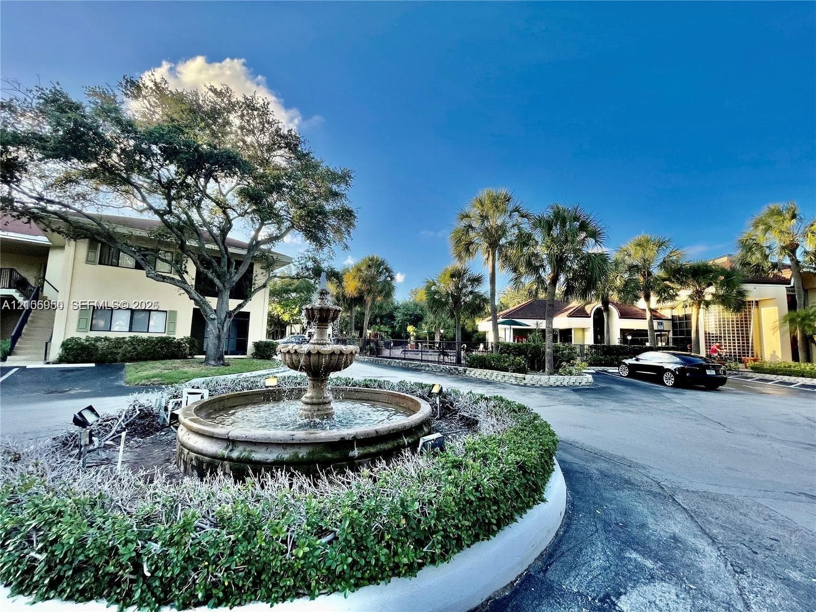 Tiered stone fountain centerpiece surrounded by manicured shrubbery, mature trees, and palm landscaping