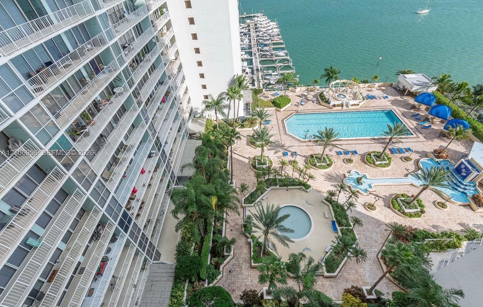 High-rise building with tiered private balconies overlooking a pool deck featuring multiple swimming pools, stone-paved landscaping, palm trees, and an attached boat marina