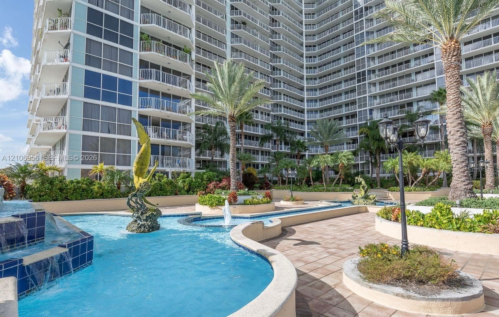 Multi-story high-rise exterior featuring a tiered pool with blue tile accents, sculptural fountains, paved walkways, and mature palm landscaping