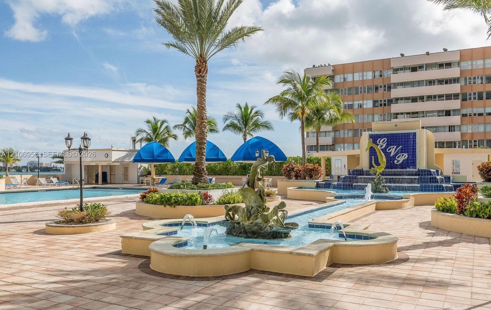 Outdoor swimming pool complex featuring decorative water fountains, paver stone hardscaping, lush palm trees, and structural shaded areas