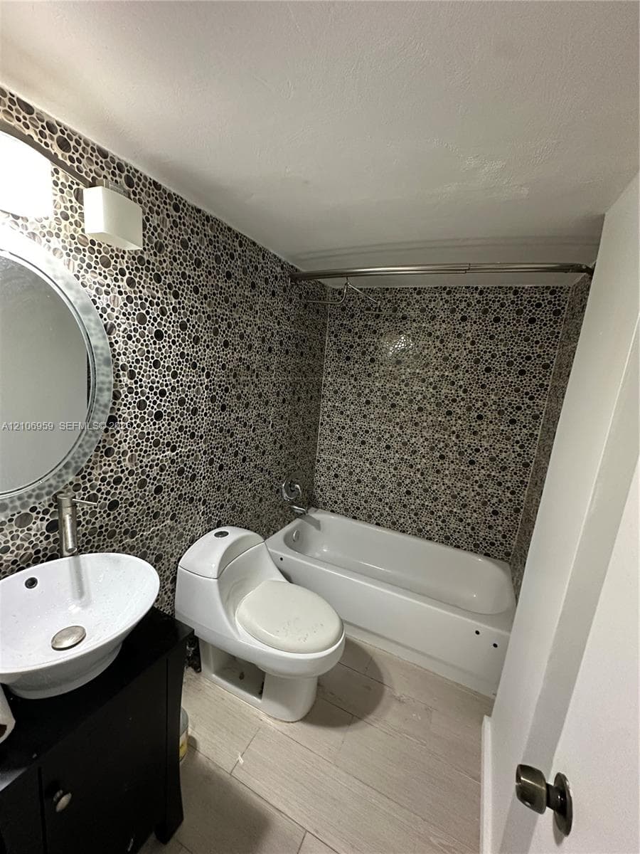 Bathroom featuring circular mosaic wall tiling, white vessel sink with a polished metal faucet, black vanity cabinet, wood-finish flooring, and a standard soaking tub