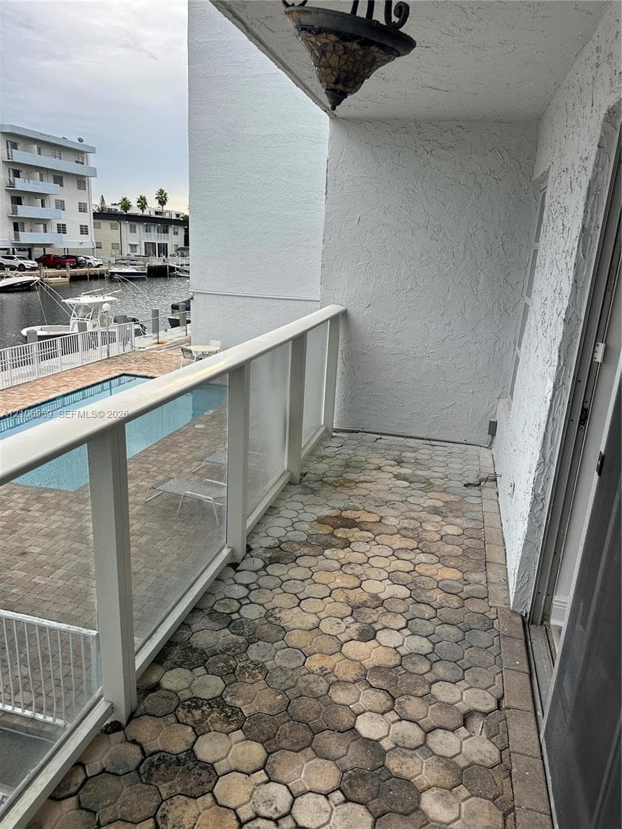 Covered balcony featuring a glass-panel railing, hexagonal stone pavers, textured stucco walls, and a decorative overhead light fixture